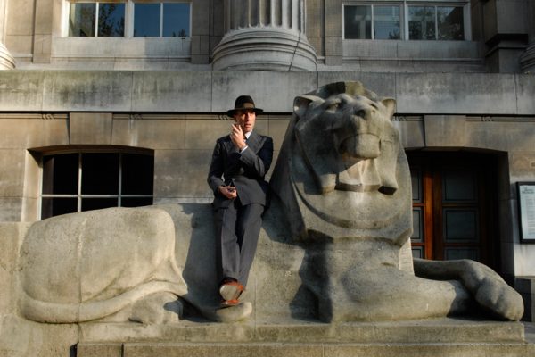 man with pipe and hat sitting on a carving of a stone lion smoking a pipe outside the British Museum